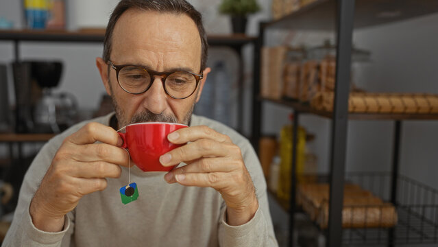 Mature man in an indoor setting enjoying a hot beverage from a red mug in a cozy mood surrounded by kitchen shelves suggesting a relaxed work break