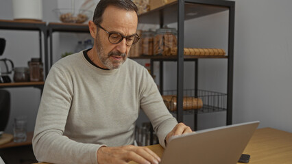 Mature man working on a laptop in an office environment, focusing intently amidst organized shelves, suggesting a professional workspace with a modern interior design.