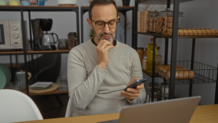 Middle-aged hispanic man pondering in a modern office room, sitting at a desk with a phone and laptop amidst stylish interior shelves and decor.