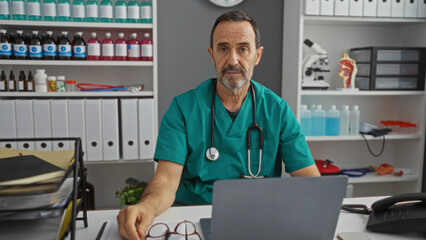 Mature hispanic man in hospital clinic setting wearing scrubs with medical equipment and documents nearby showcasing professional healthcare work environment indoors