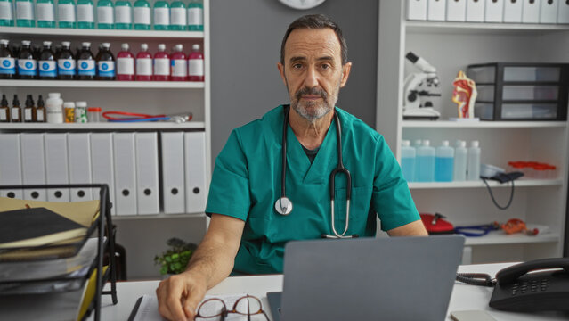 Mature hispanic man in hospital clinic setting wearing scrubs with medical equipment and documents nearby showcasing professional healthcare work environment indoors