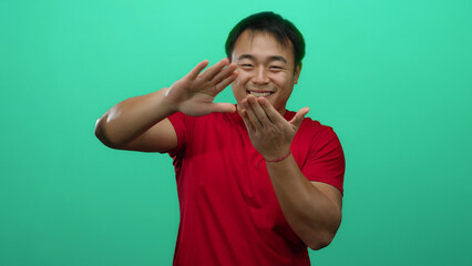 Young man in red shirt making money gesture against green background with a cheerful expression...