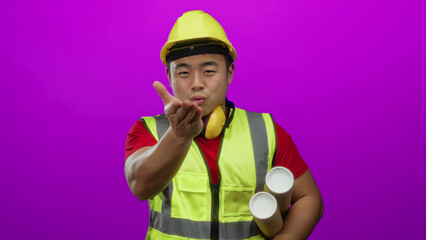 Young asian man wearing construction gear blows a kiss while holding blueprints against a vivid pink background, showcasing a playful yet professional architectural theme.