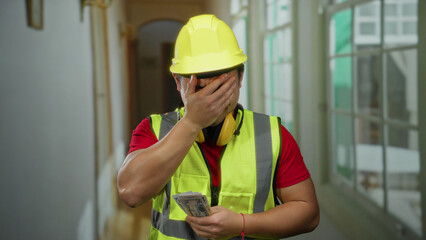 Construction worker wearing a hardhat and safety vest stands indoors distractingly holding money in a hotel hallway indicating stress or frustration.