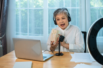 Senior influencer Asian woman presenting gift box in online live streaming with microphone and ring light