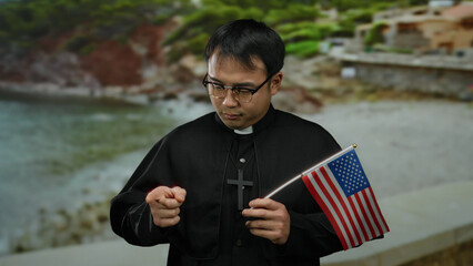 Young priest holding usa flag at seaside, engaging in thoughtful gesture with scenic beach...
