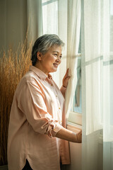 Smiling senior Asian woman looking out window with sunlight and positive mood
