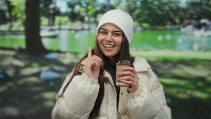Young woman in a park wearing a white coat and beanie, holding a coffee cup, smiling and pointing playfully towards the camera, surrounded by lush greenery.