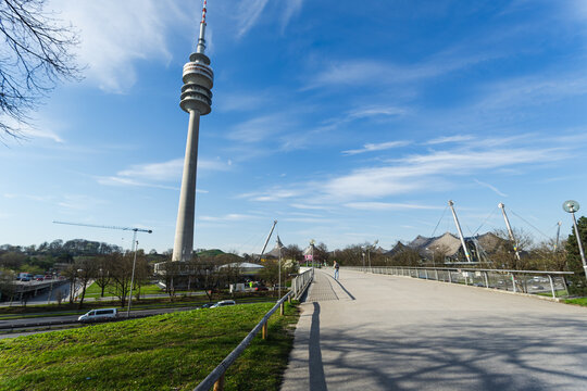 Munich, Bavaria, Germany - April 9 2025: Olympic Tower rises above Olympiapark as visitors walk along a sunlit bridge under a vibrant blue spring sky.