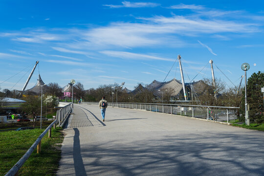 Munich, Bavaria, Germany - April 9 2025: A pedestrian walks across a bridge in Olympiapark with the iconic tent roof of the Olympic Stadium in the background on a sunny spring day.