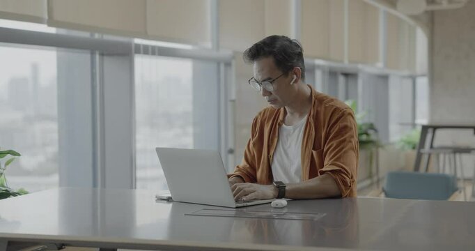 Senior Asian businessman wearing headset celebrates success during online meeting in modern co-working space. Remote work and business communication concept.	