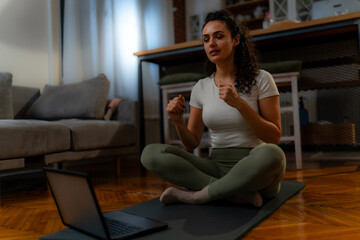 Obraz premium Young woman sitting on a yoga mat in a home environment. She using a laptop, likely engaging in an online class or activity. The woman has curly hair and is wearing casual, comfortable clothing.