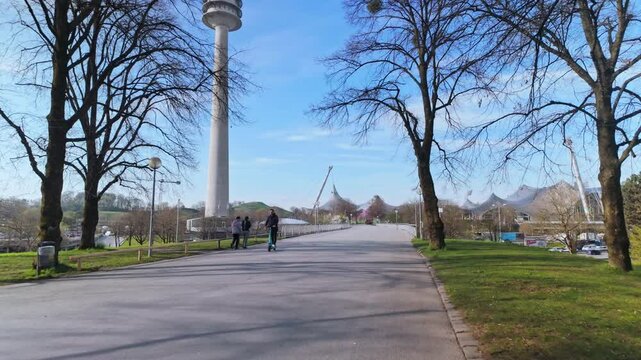 	Munich, Germany &ndash; April 9, 2025: Forward-moving shot through Olympia Park with pedestrians walking near the Olympic Tower on a clear spring day.
