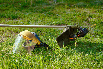 Safety mask and trimmer resting on green grass, yard care preparation, emphasizing proper protection and equipment used in outdoor lawn trimming tasks.