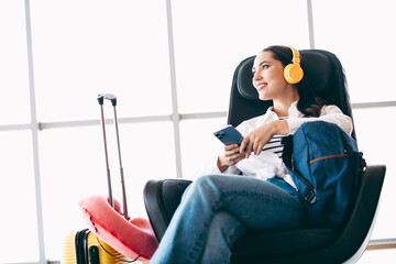 Charming woman waiting at airport with headphones enjoying music with smartphone and luggage for...