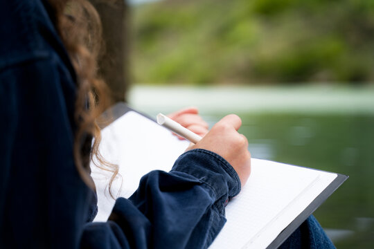 Close-up of Caucasian woman hand writing down thoughts and ideas in notebook with pen taking notes journaling keeping manifesting diary as a routine sitting in nature mountain park by lake outdoors
