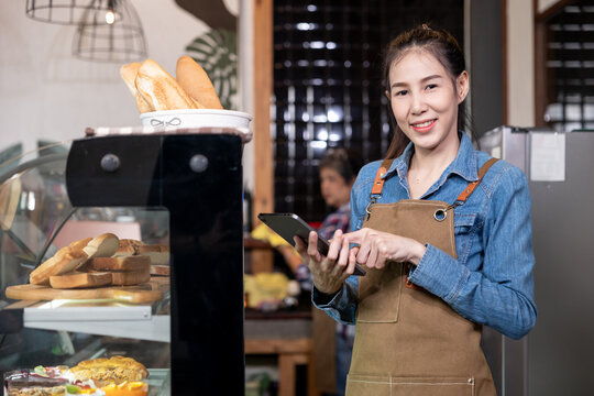 Asian young female barista wearing denim shirt and apron standing beside pastry display case smiling with confidence while holding digital tablet showing modern service and management in bakery cafe
