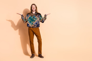 Charming young man with long hair posing playfully, showcasing an elegant outfit and confident demeanor on a beige studio background