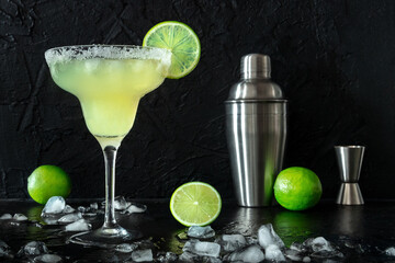 Margarita cocktail in the classic glass, side view on a black background, with ice, lime, and a salt rim, with a shaker and a jigger