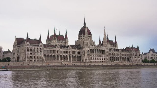 The neo Gothic Hungarian Parliament Building stands by the Danube River in Budapest, with spires and a dome visible under an overcast sky.