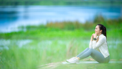 A woman is sitting on the grass by a lake. She is wearing a white shirt and blue pants
