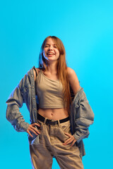 Teen girl laughs out loud in joyful motion wearing casual crop top and open denim shirt in neon light against blue studio background. Concept of happiness, human emotions, fashion, youth