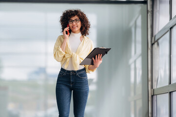 Fototapeta premium Talking by phone. Beautiful woman is standing indoors against blurred wall
