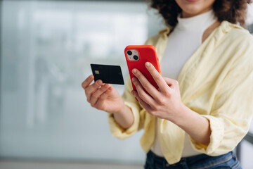 Credit card and phone in hands. Beautiful woman is standing indoors against blurred wall
