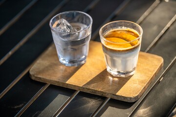 Two clear glasses of water and whiskey with ice and lemon on a wooden serving board placed on a black wooden table in a well-lit environment, close-up shot