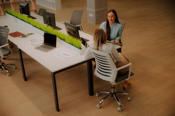 Two professional women sitting at a modern office desk, discussing work and smiling. Bright...