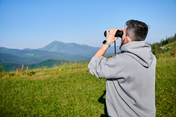 Man stands in grassy field, using binoculars to observe the distant mountains, wearing gray hoodie, smartwatch on his left wrist. Rolling green hills and clear blue sky, creating tranquil scene.