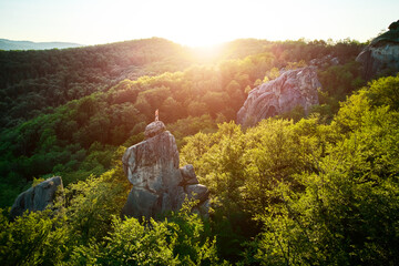 Aerial view of climber standing atop rock formation at Dovbush Rocks in Carpathian mountains, Ukraine. Sun sets, casting warm glow over lush, green forest, distant hills, creating breathtaking view.