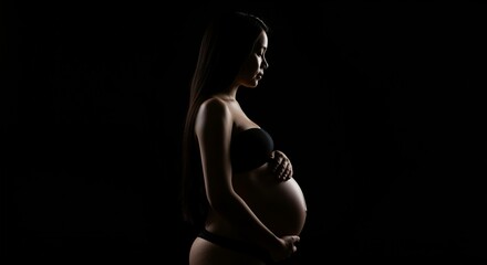 Silhouette of pregnant woman in dark setting, hands gently cradling her belly in a gesture of love.