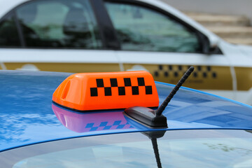 Close-up of a blue taxi car with orange checkered symbol on hood, another taxi in background, urban transportation service.