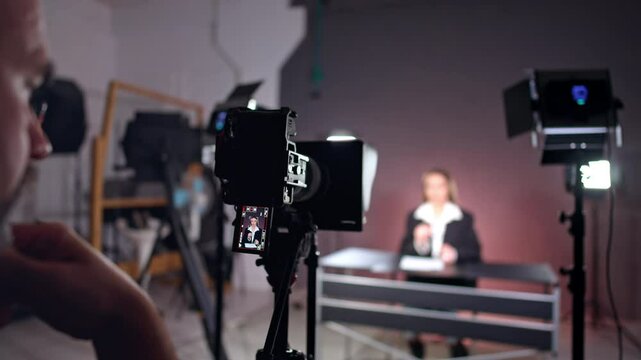 Backstage of the photo studio work. Male cameraman sits at camera recoding video of a female reporter.