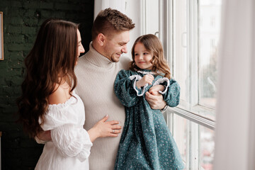 Family day. Happy young mother and father with love embrace little child daughter by window at home, stylish parents in festive clothes hold hands small kid relax rest in living room. Children's day.