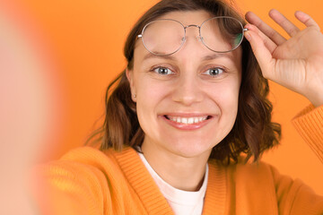 Playful young woman takes a selfie, smiles, sticks out her tongue, holds glasses on yellow backdrop. Modern lifestyle, social media, youth influencer marketing, self expression, personal branding