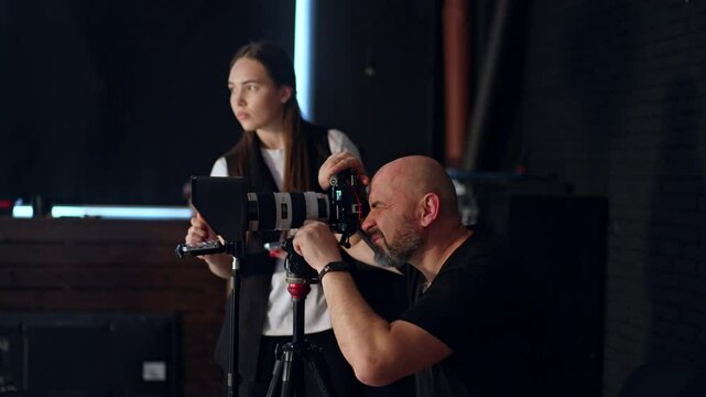 Bald bearded photographer looks at camera on tripod. Girl at backdrop uses a smartphone. Photo studio footage backstage.