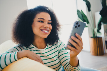 Young woman with curly hair smiling at her smartphone while relaxing on a cozy couch in a bright modern living room