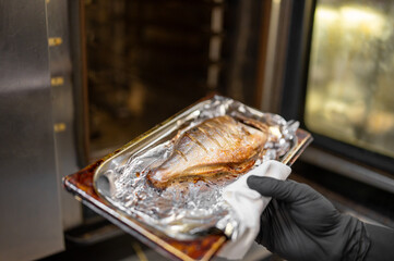 A gloved hand holds a tray with a baked fish covered in foil, being removed from an oven. The fish...