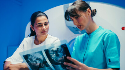 Positive female patient listening carefully while medical worker in teal outfit explaining brain scan results. Focused technician pointing at radiograph during post-MRI discussion near tomography unit