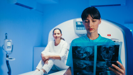 Concentrated female doctor analyzing skull scan while patient sitting behind on tomography bed...