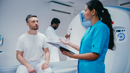 Male patient sitting on MRI table explaining issue, gesturing with lips pursed. Female nurse in blue scrubs listening, holding clipboard. Doctor adjusting MRI machine behind in blurred background.