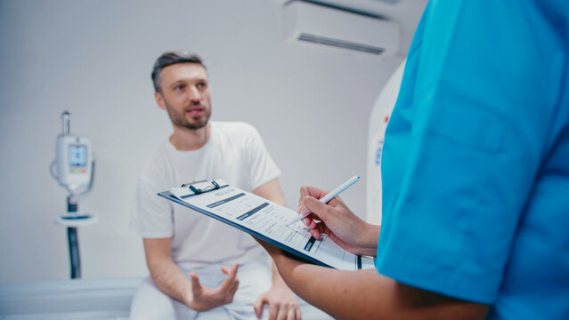Concerned Caucasian man explaining condition while pointing at body and sitting on examination table. Medical worker in blue uniform writing and documenting information during patient examination.