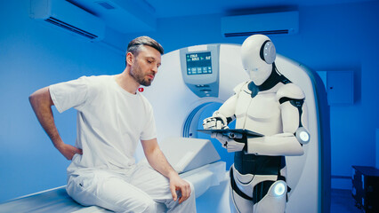 Worried Caucasian man touching lower back and describing pain while talking to futuristic robot. Advanced humanoid assistant analyzing symptoms and entering data on tablet before conducting scan.