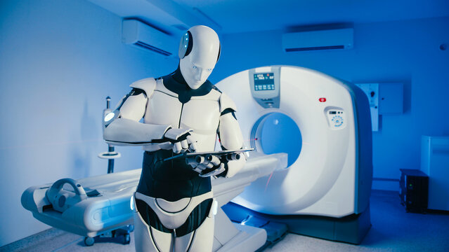 Humanoid robot operating touchscreen tablet while standing beside advanced CT scanner in clinical room. Machine processing patient data or controlling diagnostic system during autonomous procedure.