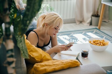 Teenage girl relaxing on sofa using smartphone with snacks and notebook nearby
