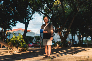 Middle aged Southeast Asian man with arm in sling walking in a park, showing resilience and...