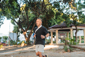 Middle aged Southeast Asian man jogs at a fast pace wearing an arm sling, demonstrating grit,...