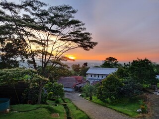 Beautiful sunset view over a green countryside with lush trees and simple village houses. The golden-orange sky blends harmoniously with the silhouettes of the trees,